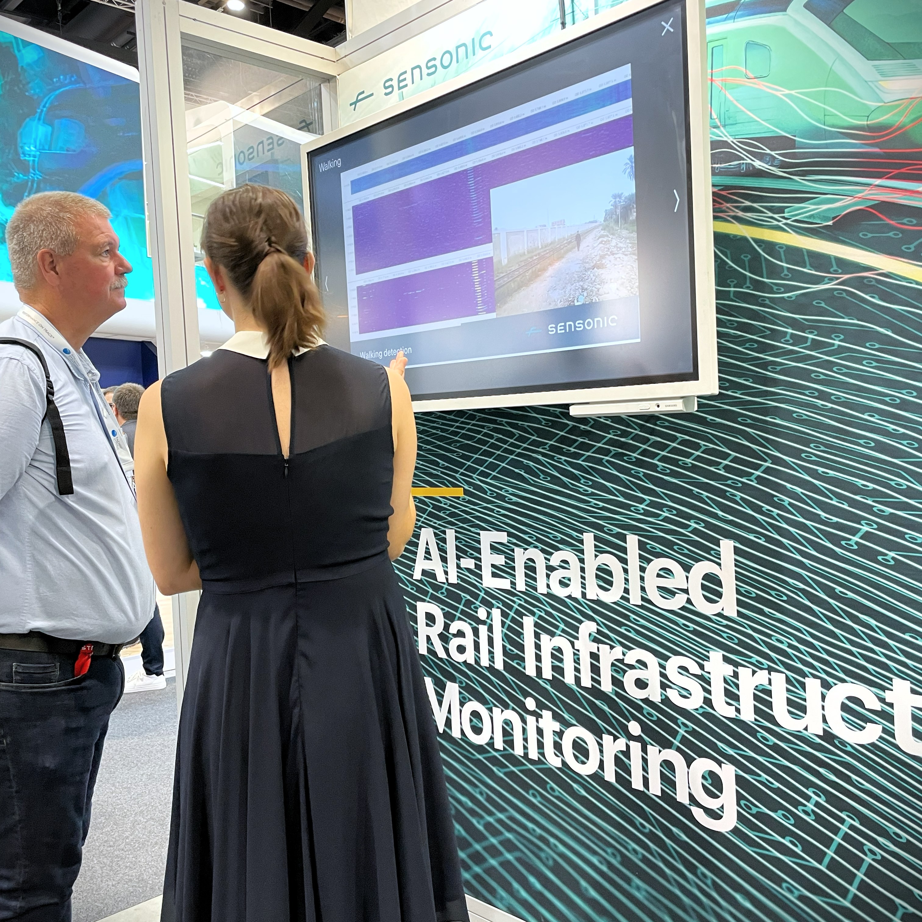 Lady talking to a man infront of s TV screen. Text below the screen reads AI-Enabled rail infrastructure monitoring